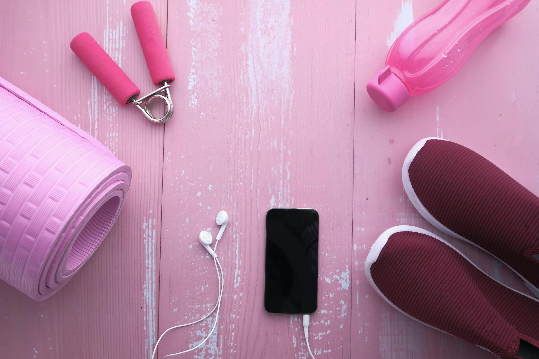 Akcesoria fitness na różowym tle: mata, buty sportowe, woda i smartfon